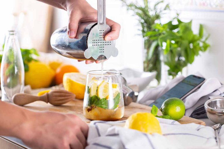 Mixologist pouring an alcoholic drink in a cup while watching an instructional video on phone at home.