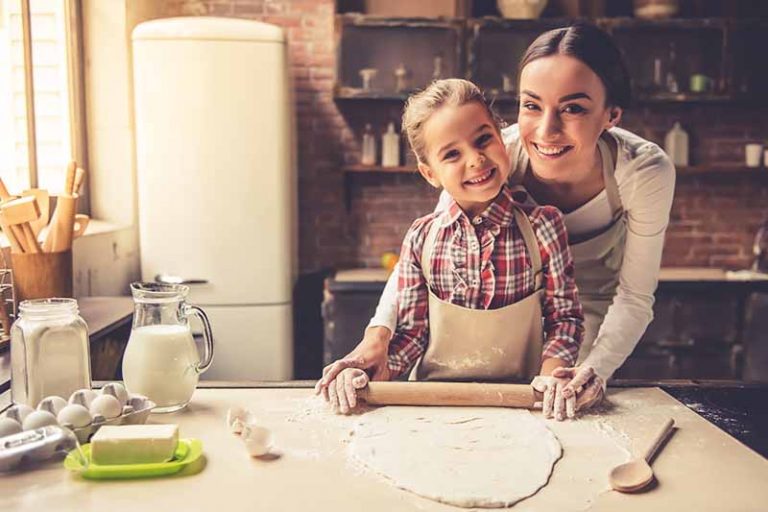 Horizontal image of a mother and daughter smiling as they roll out pastry on the counter.