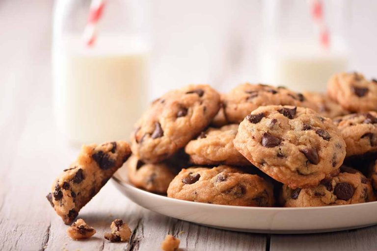 Horizontal image of a plateful of chocolate chip cookies on a white wooden table in front of milk bottles.