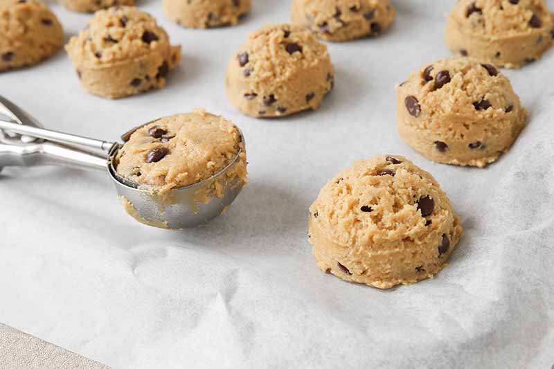 Horizontal image of scoopfuls of unbaked dough in rows on a baking sheet lined with parchment paper.