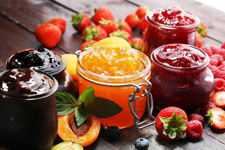 Horizontal image of various jams and jellies in glasses on a wooden table surrounded by fresh produce.