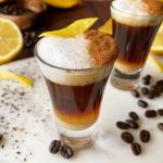 Horizontal image of two shooters filled with a layered espresso beverage topped with frothed milk, cinnamon, and lemon peel.