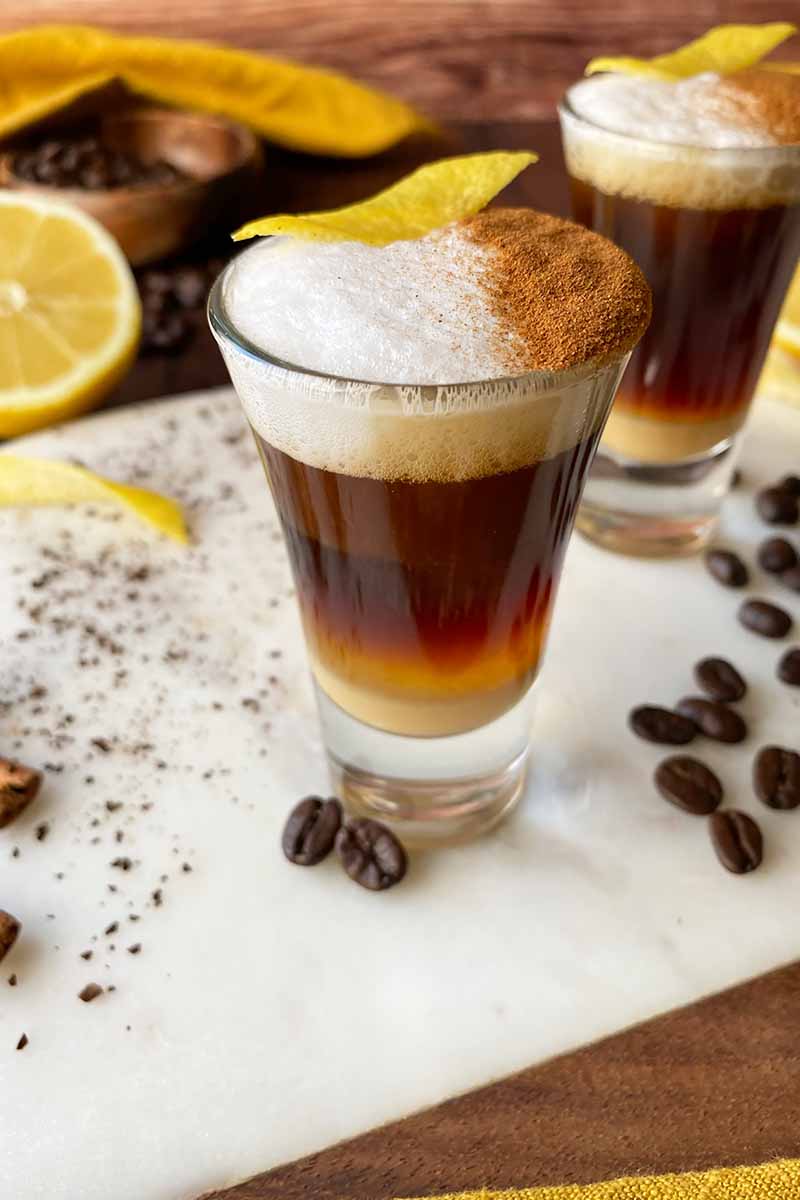 Vertical image of two shooters filled with a layered espresso beverage topped with frothed milk, cinnamon, and lemon peel.