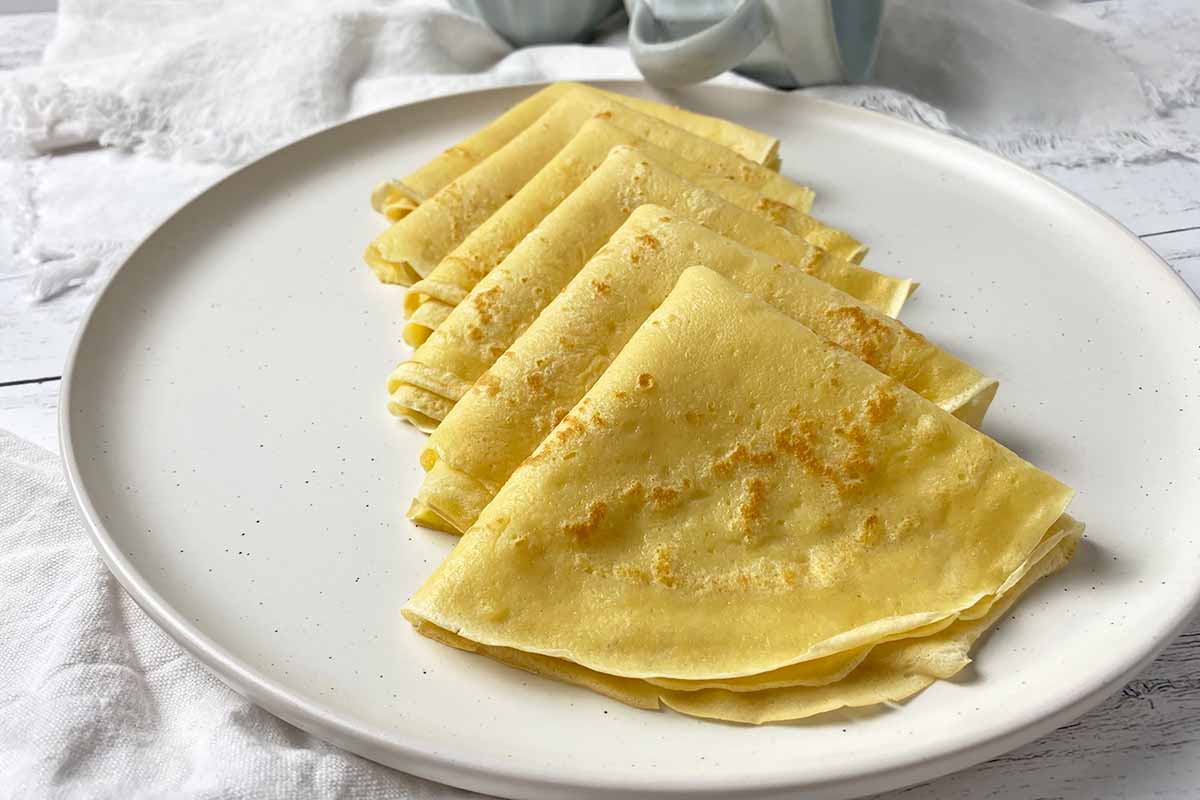 Horizontal image of a row of folded thin pancakes on a white plate surrounded by white towels, with mugs in the background.