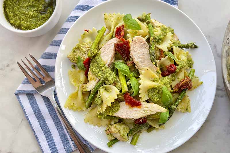 Horizontal image of a white dinner plate with farfalle mixed with chicken slices and assorted vegetables in a green sauce next to a fork and napkin.