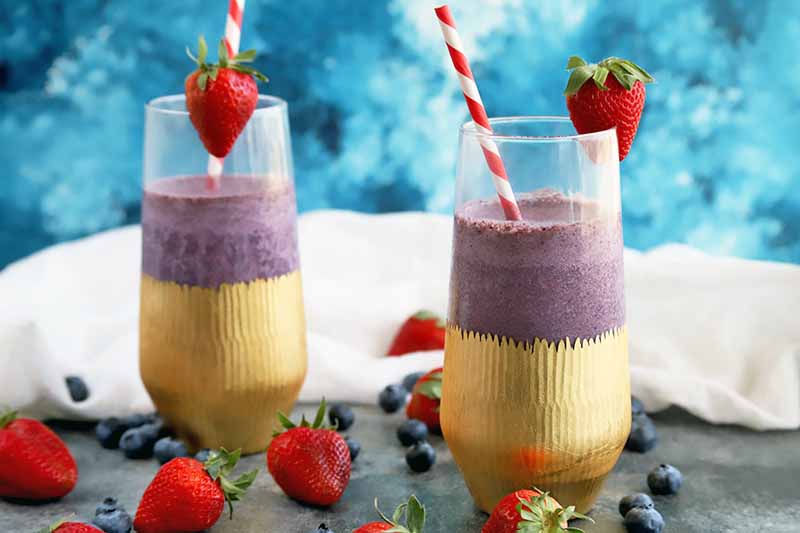Horizontal image of a purple smoothie in two tall glasses with gold paper wrappers at the base of each, with red and white striped paper straws and a whole strawberry garnish, with scattered berries on a gray surface with a white cloth in the background, against a mottled blue and white backdrop.