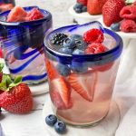 Horizontal close-up image of blue-rimmed glassware filled with a light pink liquid, ice cubes, and assorted fresh garnishes on white coasters in front of white cake stands.