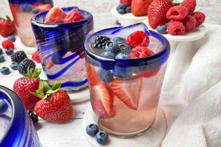 Horizontal close-up image of blue-rimmed glassware filled with a light pink liquid, ice cubes, and assorted fresh garnishes on white coasters in front of white cake stands.