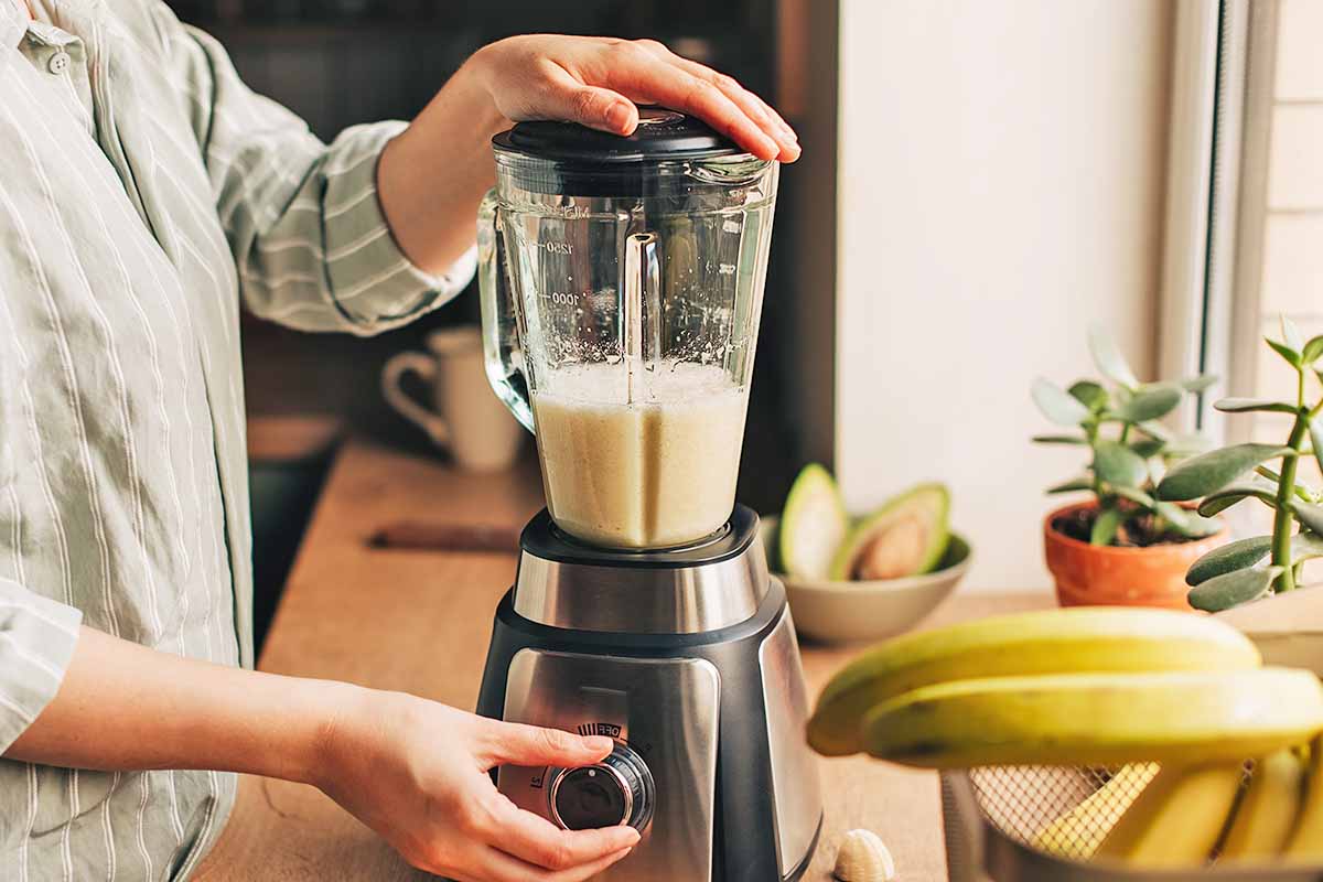 Horizontal image of a woman preparing a shake on a counter next to plants, bananas, and avocados.