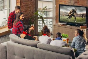 Horizontal image of an excited group of friends watching football at home while eating assorted food and drinking beer.