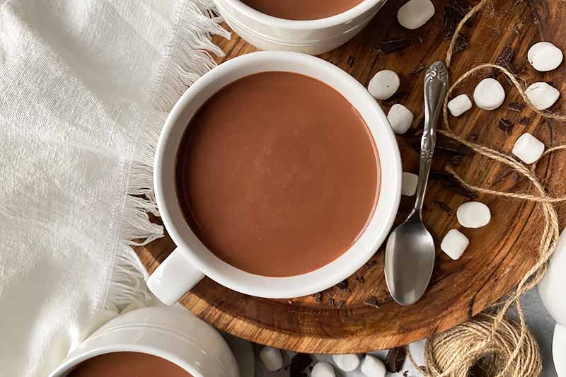 Horizontal image of mugs of a creamy brown beverage next to spoons, marshmallows, and a white towel, on a wooden board.
