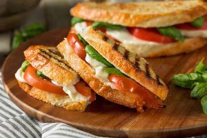 Horizontal close-up image of a sliced grilled basil, mozzarella, and tomato sandwich on a wooden cutting board on a towel next to fresh herbs.
