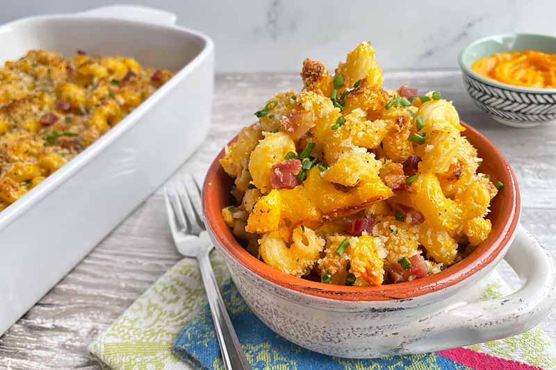 Horizontal image of a small bowlful next to a large white casserole dish of macaroni and cheese.