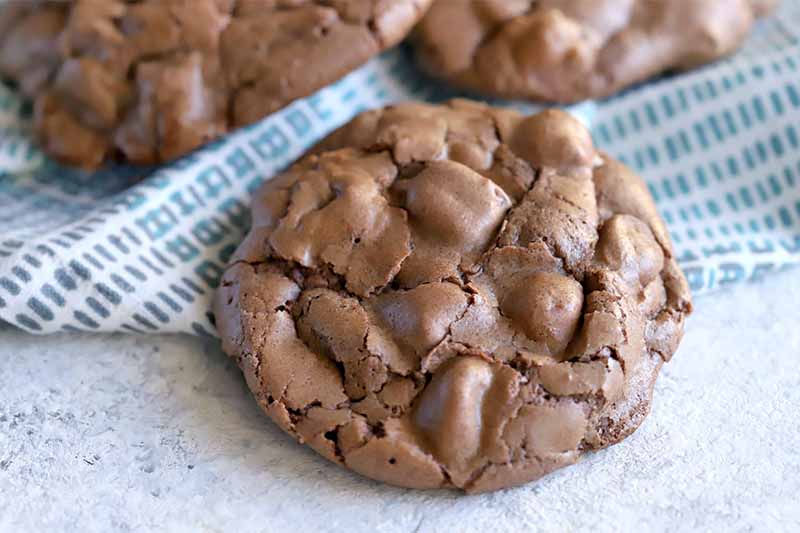 A chocolate cookie is nearly centered in the foreground with more in the background mostly beyond the edge of the frame, on a blue cloth on top of a speckled white and blue surface.
