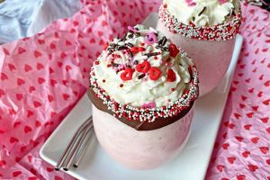 Horizontal image of two glasses with a creamy pink beverage garnished with whipped cream, chocolate rim, and sprinkles on a white plate.