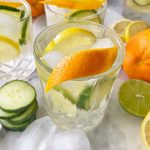 Horizontal image of glasses filled with ice, a clear liquid, and slices of fresh produce, with a thin rind as garnish.