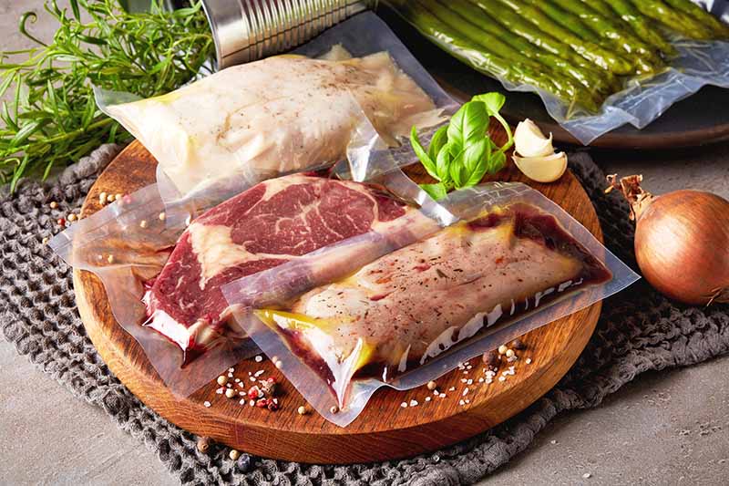 Horizontal image of sealed cuts of raw protein on a cutting board next to herbs and vegetables.