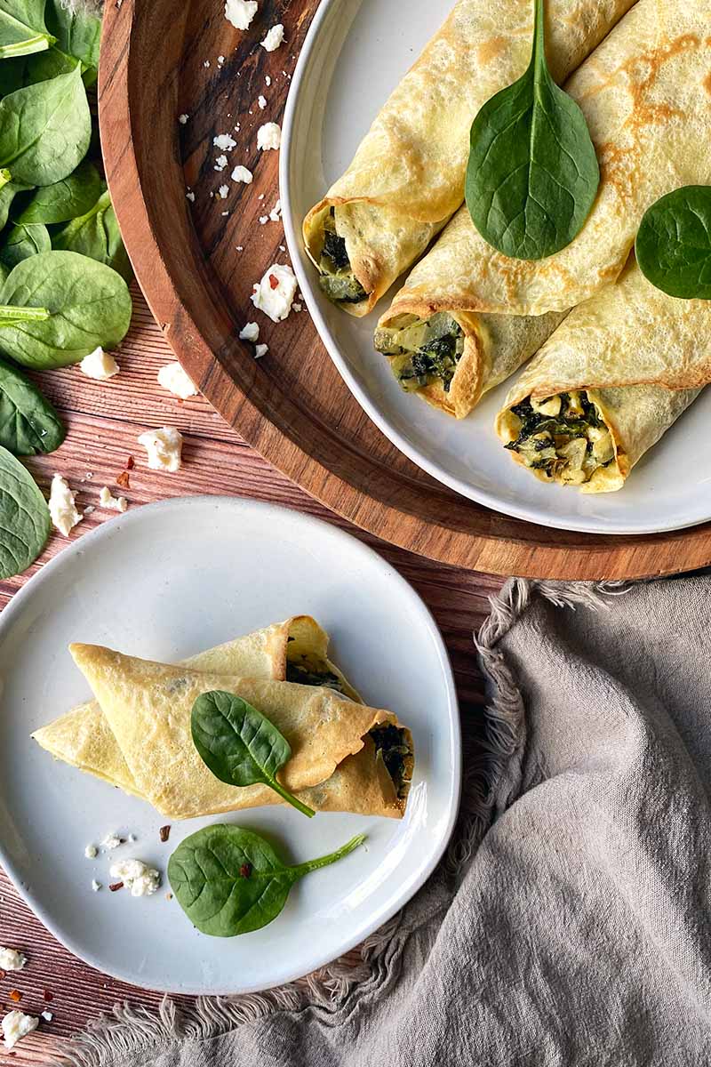 Vertical top-down image of rolled French thin pancakes with a greens and onion stuffing on a serving platter and white plates.
