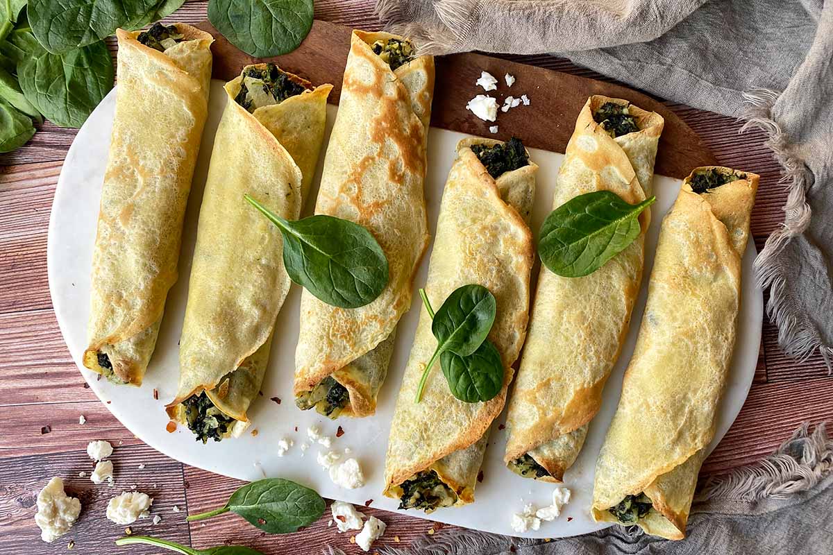 Horizontal top-down image of rolled French thin pancakes with a greens and onion stuffing on a serving platter.