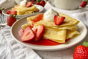 Horizontal image of two white plates, each with with a stack of folded thin pancakes served with whipped cream, sliced fruit, and a red syrup.