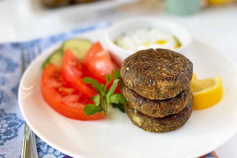 Horizontal image of a stack of garbanzo bean patties on a white platter with tomato slices, dip, lemon, and cucumbers.