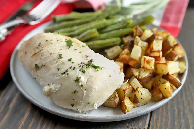 Horizontal image of a slow cooked herb chicken breast on a plate with portions of green beans and roasted potatoes, on a brown wood table with red cloth napkins and silverware in the background.