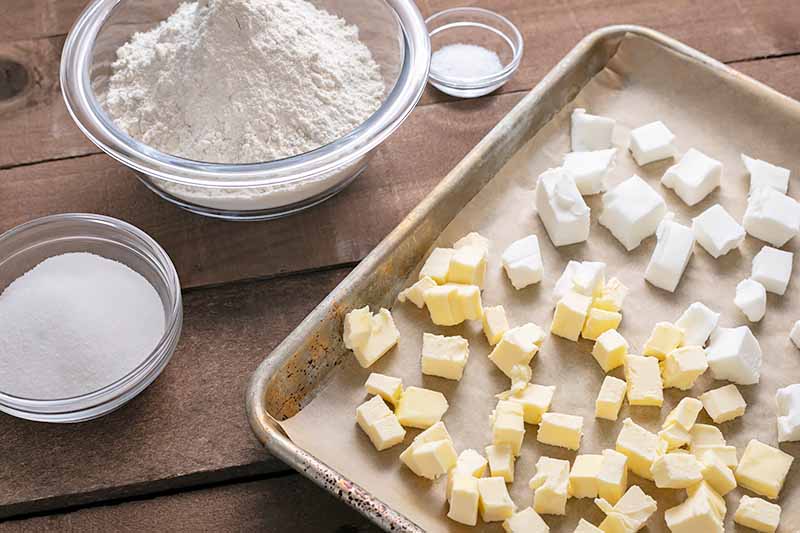 Horizontal image of a baking sheet with cubes of butter and shortening next to other measured ingredients in bowls.