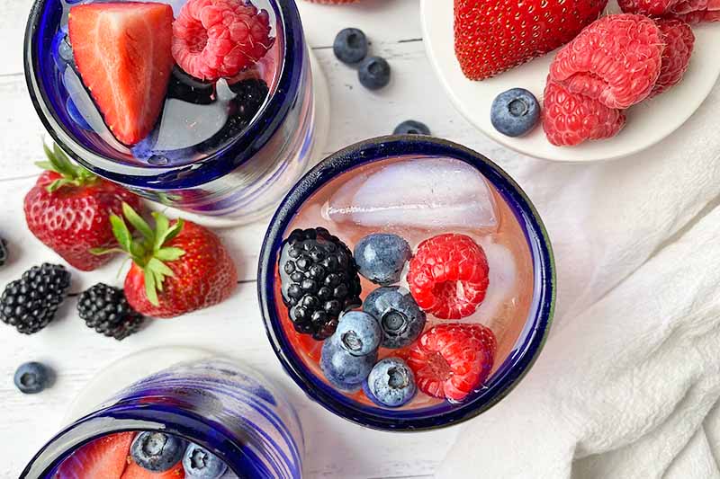 Horizontal top-down image of blue-rimmed glassware filled with a light pink liquid, ice cubes, and assorted fresh garnishes on white coasters in front of white cake stands.