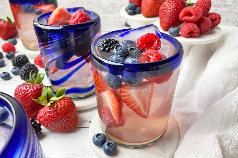 Horizontal close-up image of blue-rimmed glassware filled with a light pink liquid, ice cubes, and assorted fresh garnishes on white coasters in front of white cake stands.