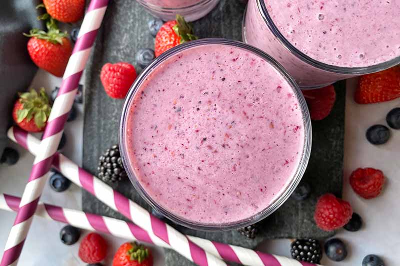 Horizontal top-down image of cups filled with a bright purple shake next to fresh fruit and straws.