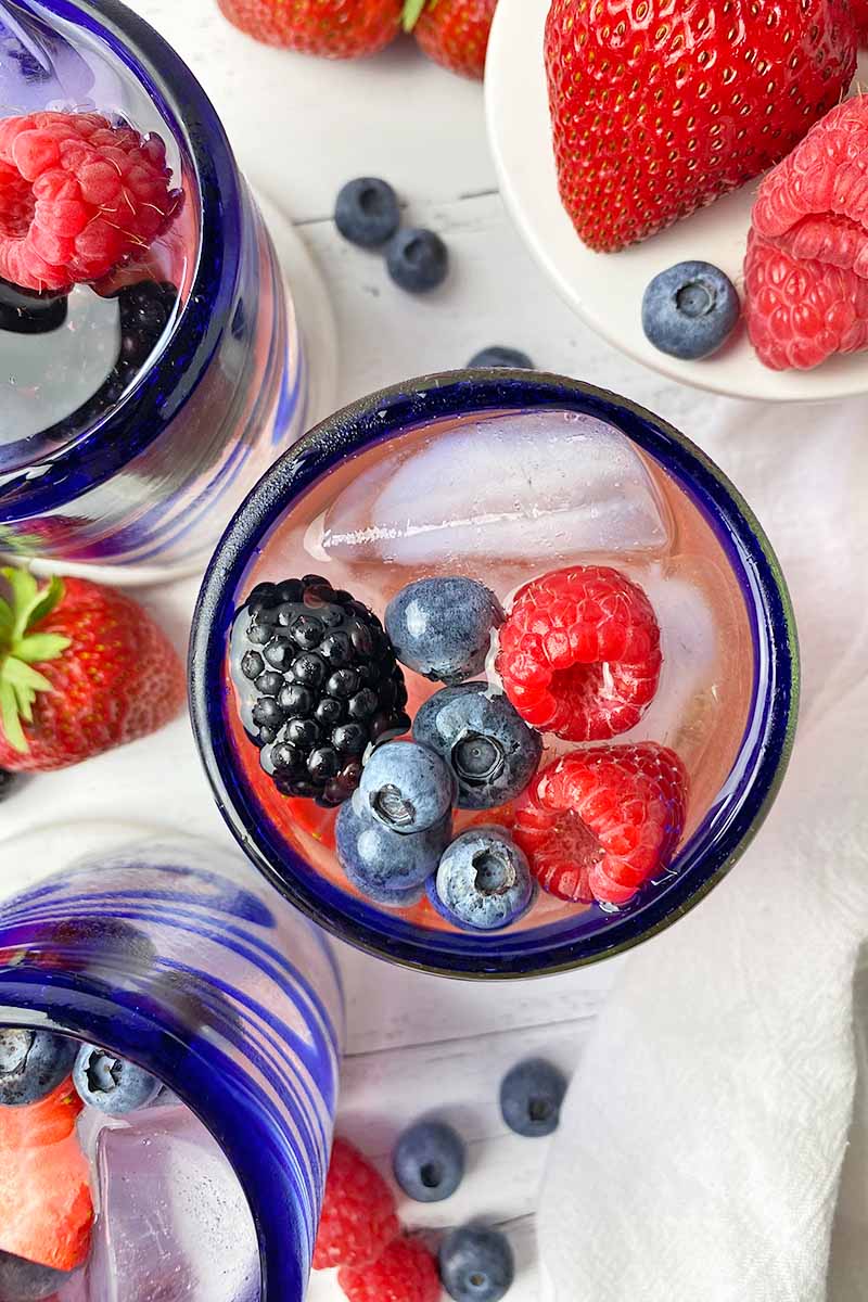 Vertical top-down image of three blue-rimmed glasses filled with a light pink beverage, ice cubes, and assorted fresh garnishes.