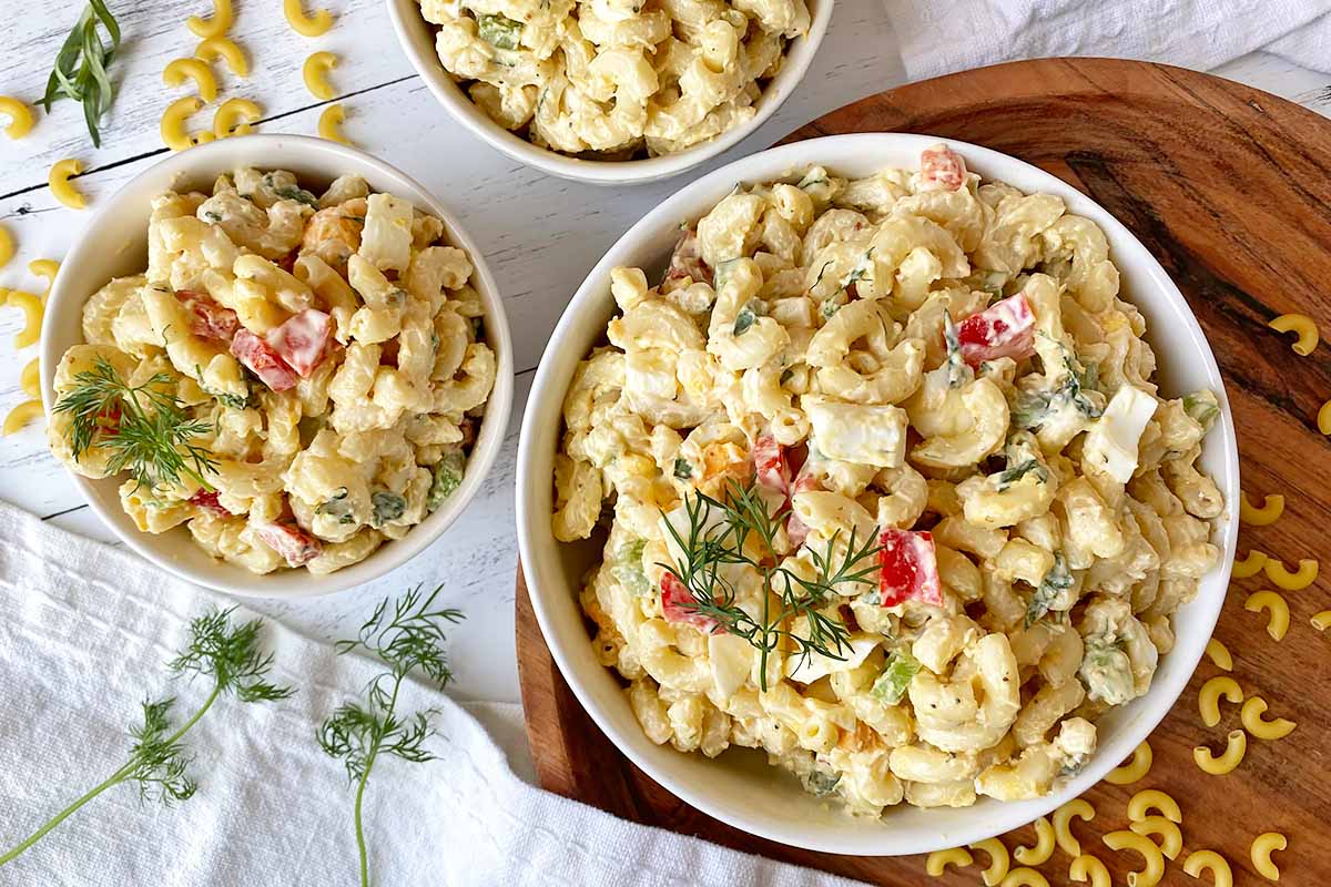 Horizontal top-down of a large bowl and two small bowls filled with a chilled macaroni dish garnished with dill fronds.