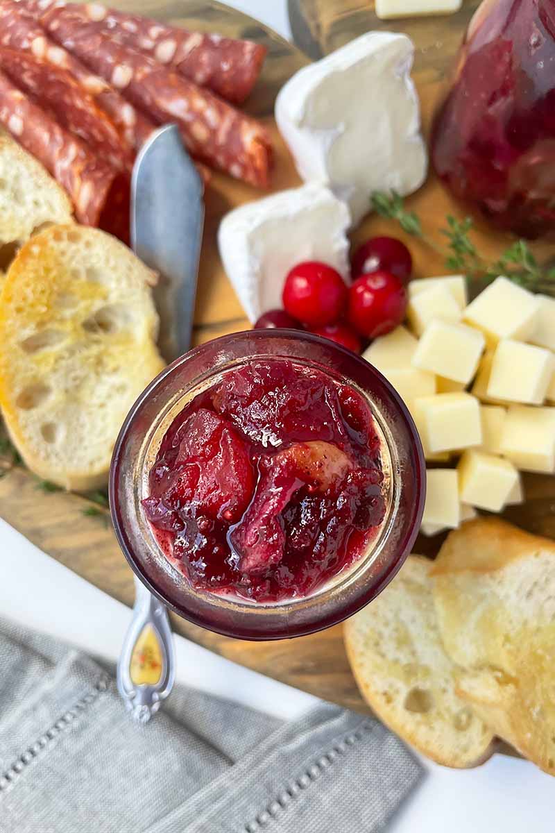 Vertical image of a jar of chunky fruit spread surrounded by items on a cheese board.