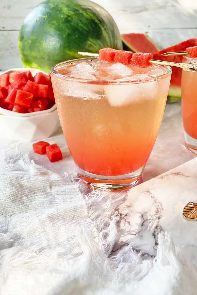Vertical image of a beverage with a beautiful pink ombre effect, next to a white towel and fresh fruit cubes in a bowl.