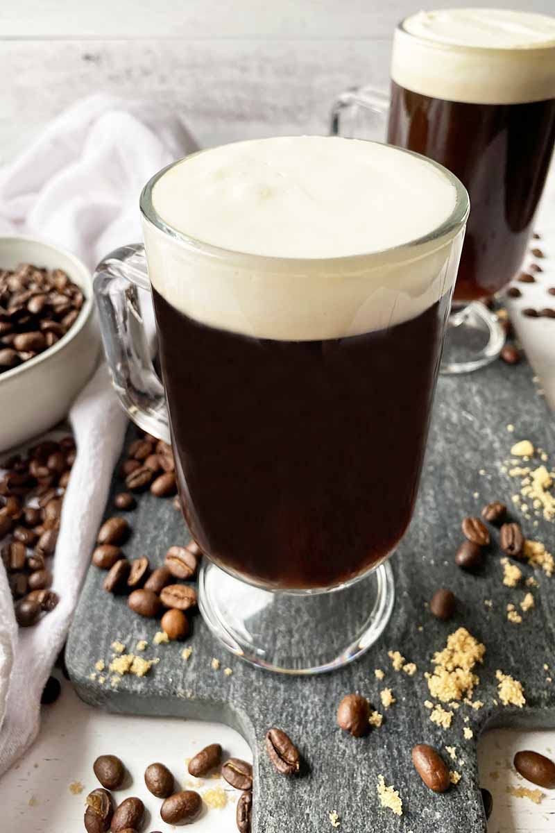 Vertical image of two mugs filled with a deep brown beverage with a fluffy white topping on a gray slate.
