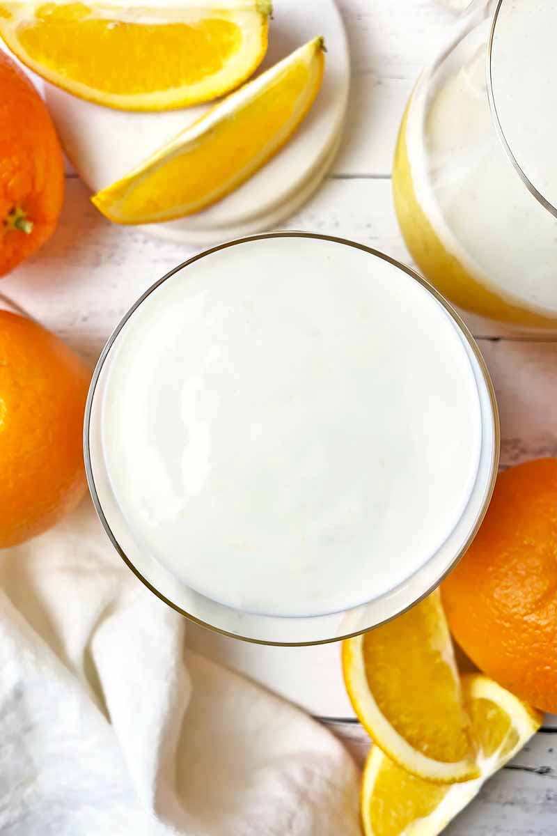 Vertical top-down image of glasses filled with a vanilla and orange beverage surrounded by sliced fruit.