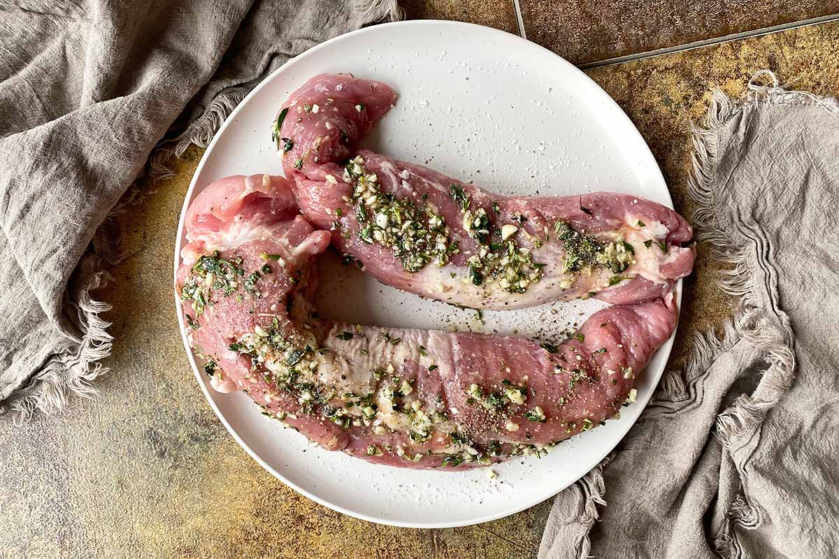 Horizontal image of two marinated pieces of raw meat on a white plate next to tan towels.