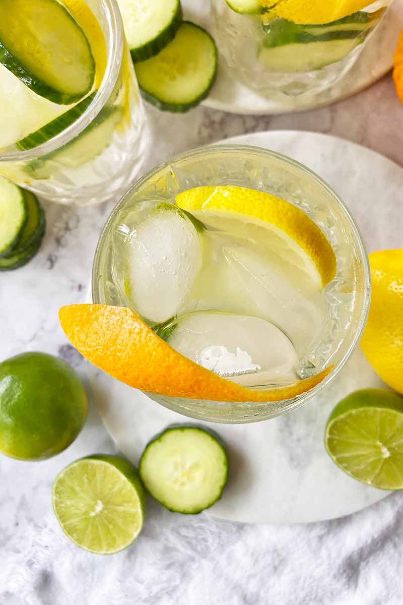 Vertical top-down image of glasses filled with a beverage, ice cubes, slices of fresh produce, and a piece of rind as garnish.