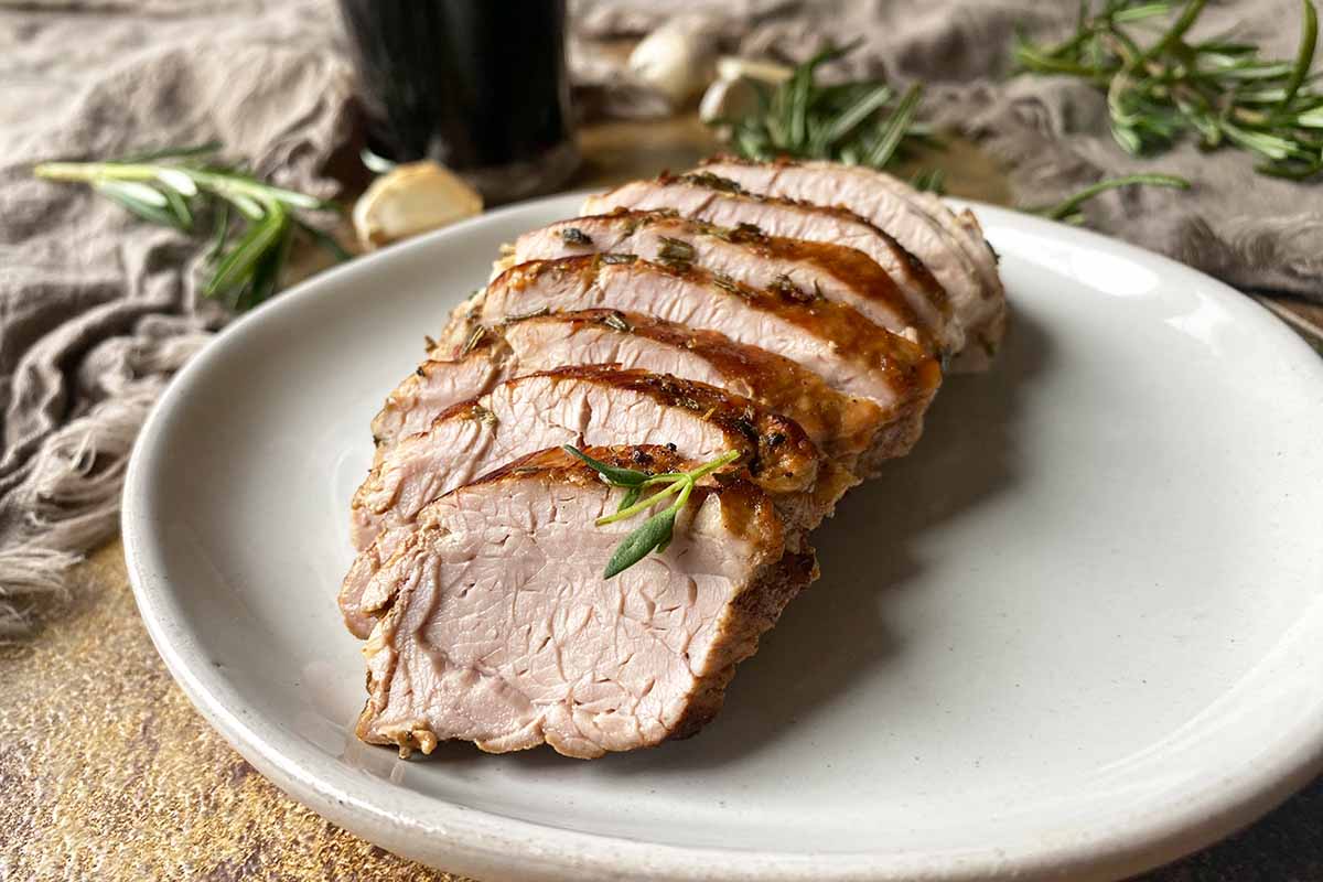 Horizontal image of slices of seared meat shingled on a white plate next to tan towels, herbs, and a glass of stout.