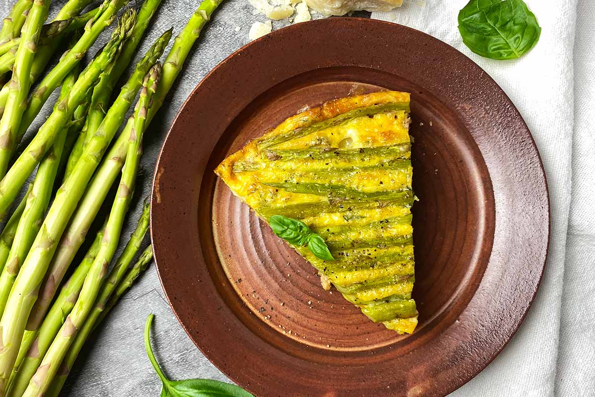 Horizontal top-down image of a wedge of a vegetable and meat frittata garnished with basil on a brown plate.