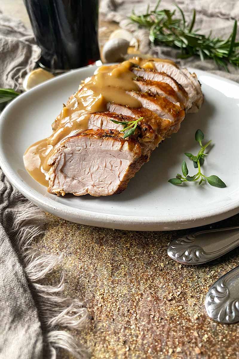 Vertical image of singled seared slices of meat half-covered in gravy on a white plate next to thyme leaves, with a glass of beer and herbs in the background.