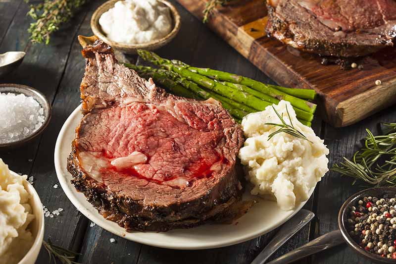 Horizontal image of a slice of rare meat on a plate with mashed potatoes and asparagus next to sauces.