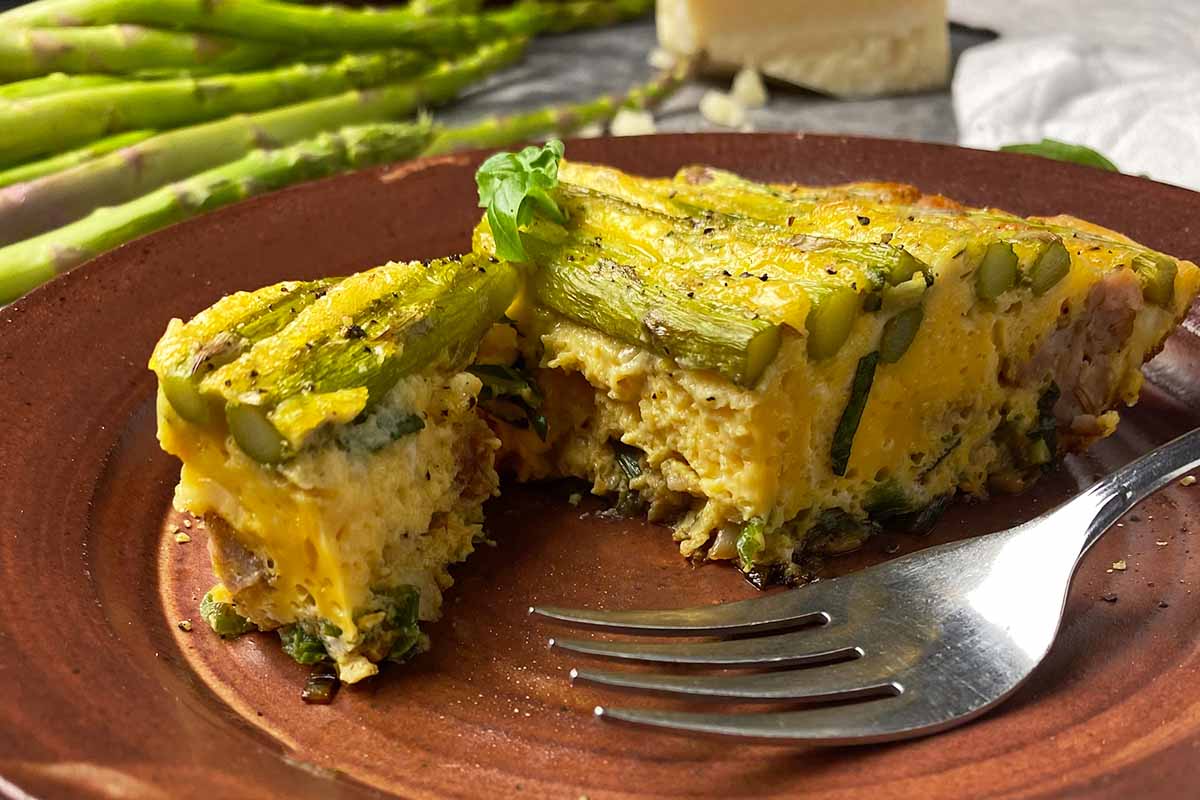 Horizontal close-up image of cutting into a wedge of a savory breakfast dish topped with vegetables on a brown plate.