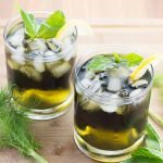 Horizontal image of two glasses with a green liquid topped with ice, mint, and lemon next to green fronds on a light wooden surface.