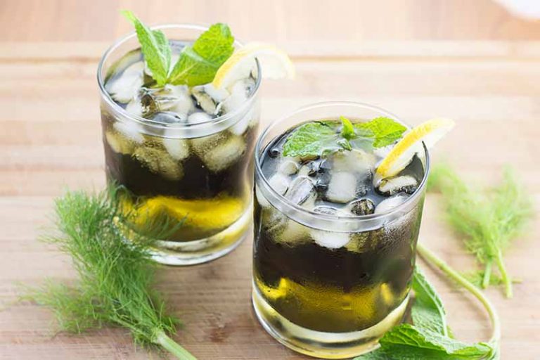 Horizontal image of two glasses with a green liquid topped with ice, mint, and lemon next to green fronds on a light wooden surface.