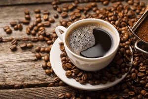 Horizontal image of a mug filled with a hot java beverage on a small plate next to beans on a wooden table.