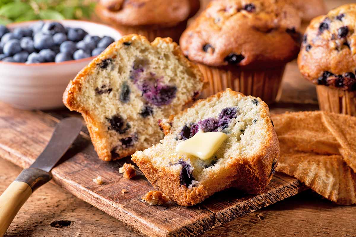 Horizontal image of a fresh blueberry pastry cut in half with butter spread on one side, on a wooden platter next to a bowl of fresh blueberries.