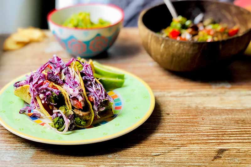 Horizontal image of a green plate with three corn tortillas filled with slaw and other assorted vegetables next to two bowls.