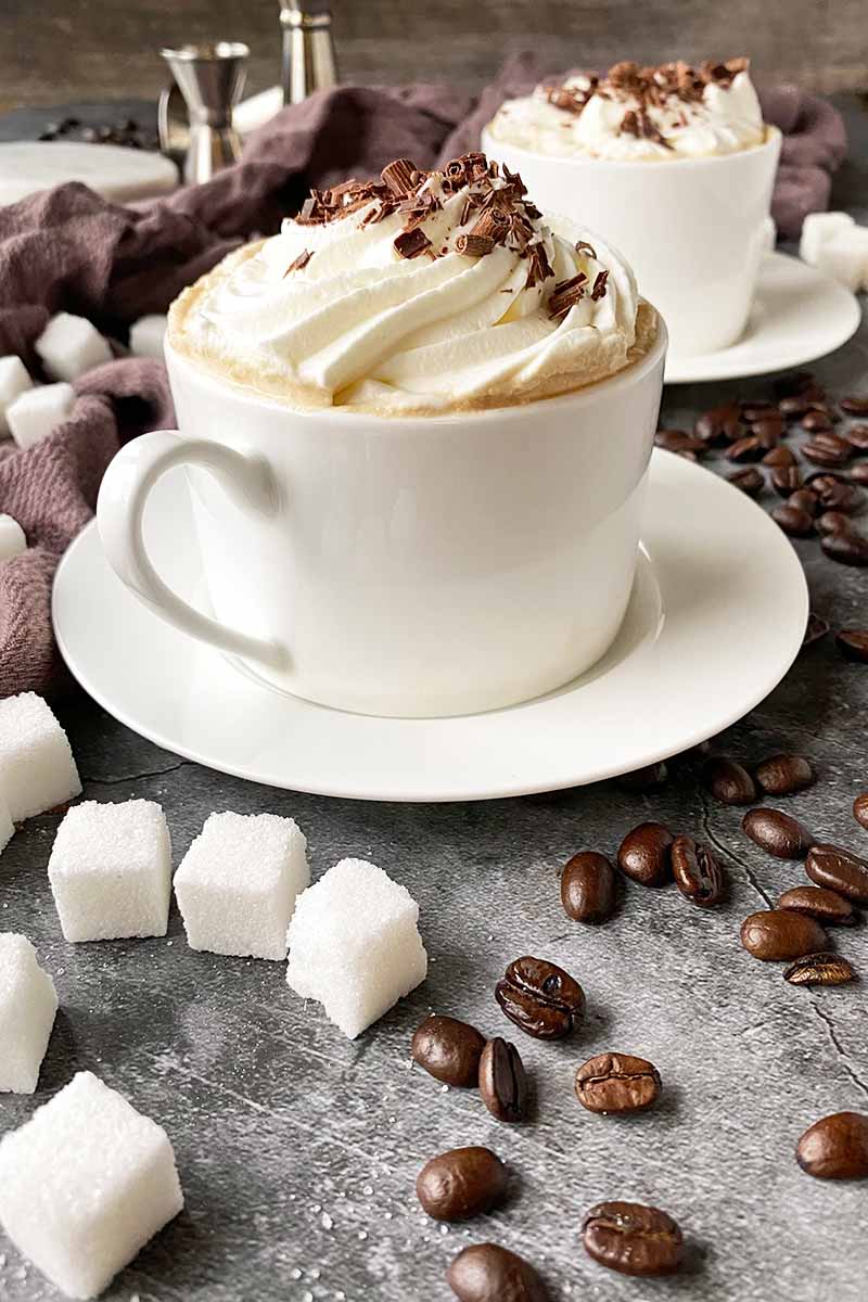 Vertical image of two white mugs filled with a cocktail topped with garnishes and chocolate shavings next to sugar cubes.