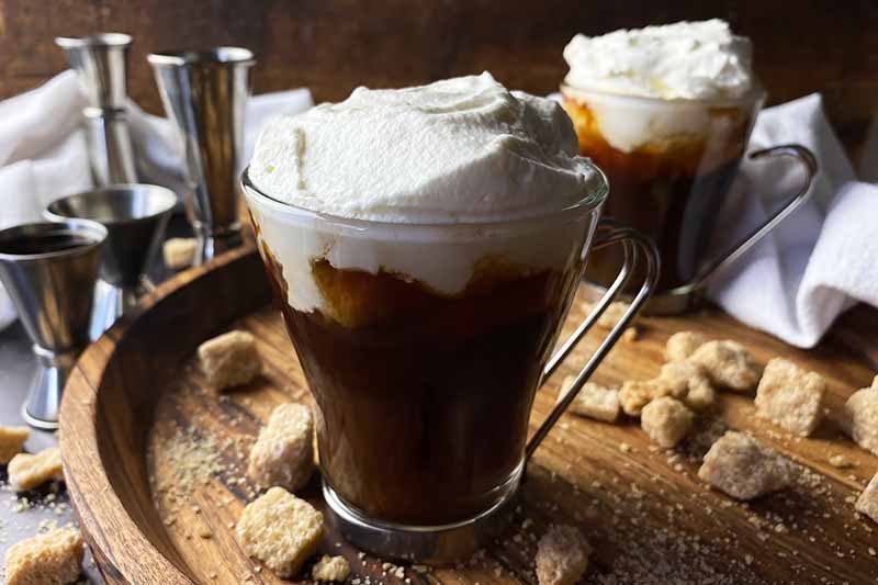 Horizontal image of a glass mug filled with a dark brown drink topped with a white foam on a wooden board with sugar cubes.